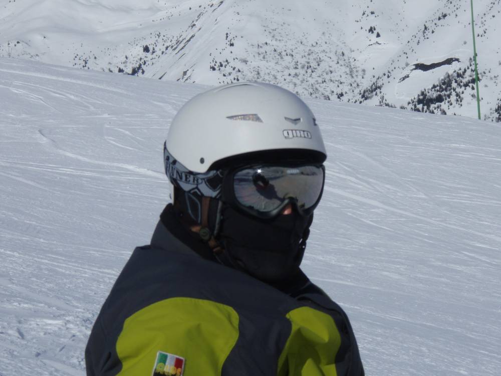 A close-up portrait of a snowboarder wearing a helmet, goggles and a balaclava against a snowy backdrop.