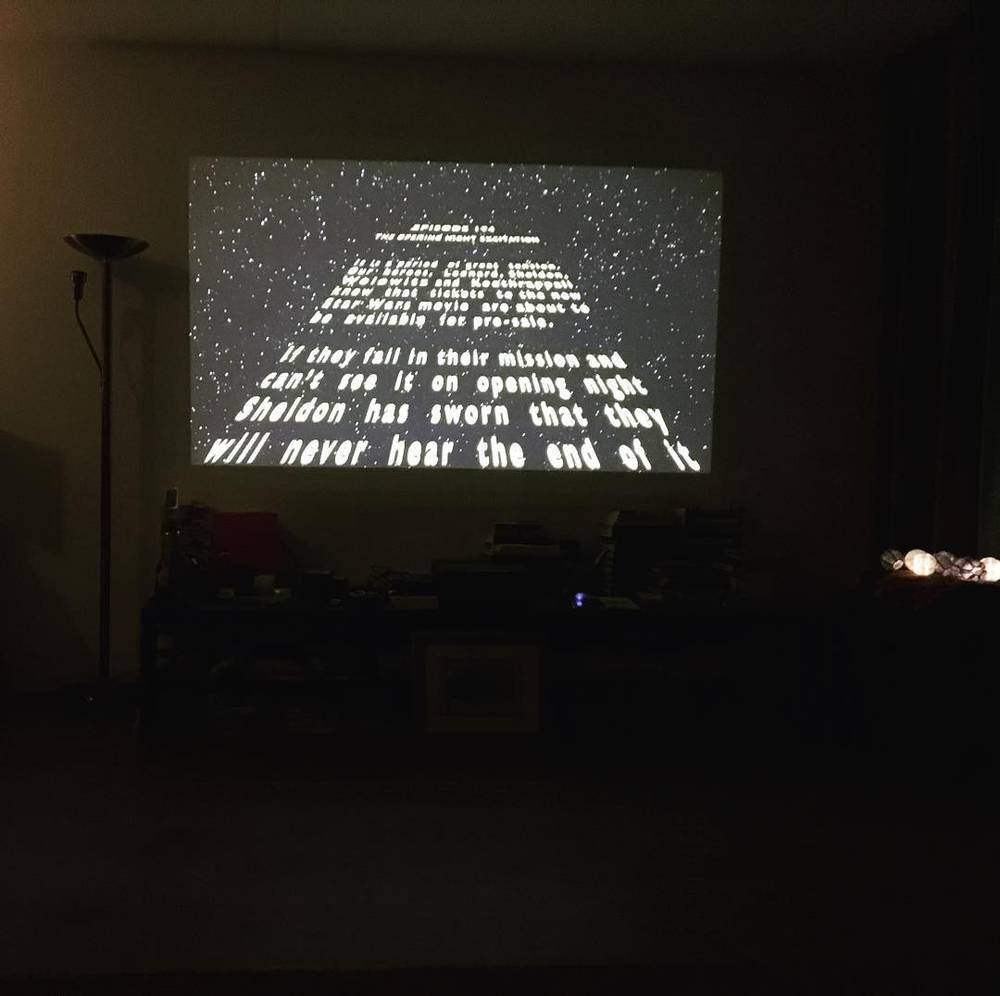 A dark room with a projector displaying scrolling text, styled like a Star Wars opening crawl referencing The Big Bang Theory.