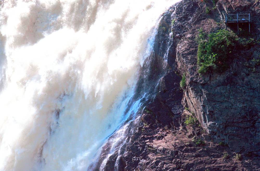 A massive, roaring waterfall rushes down a rocky cliffside, with a tiny wooden viewing platform perched near the top.