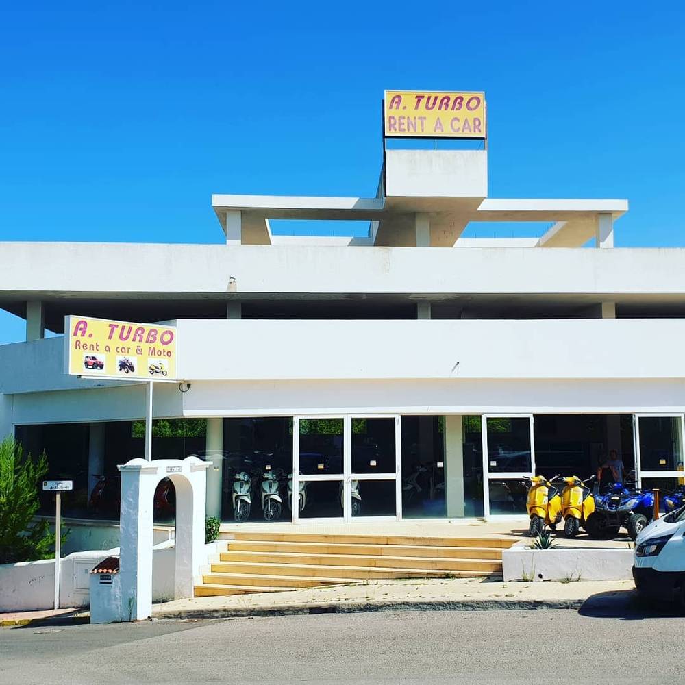 The exterior of an 'A. Turbo Rent a Car' building with mopeds parked outside.