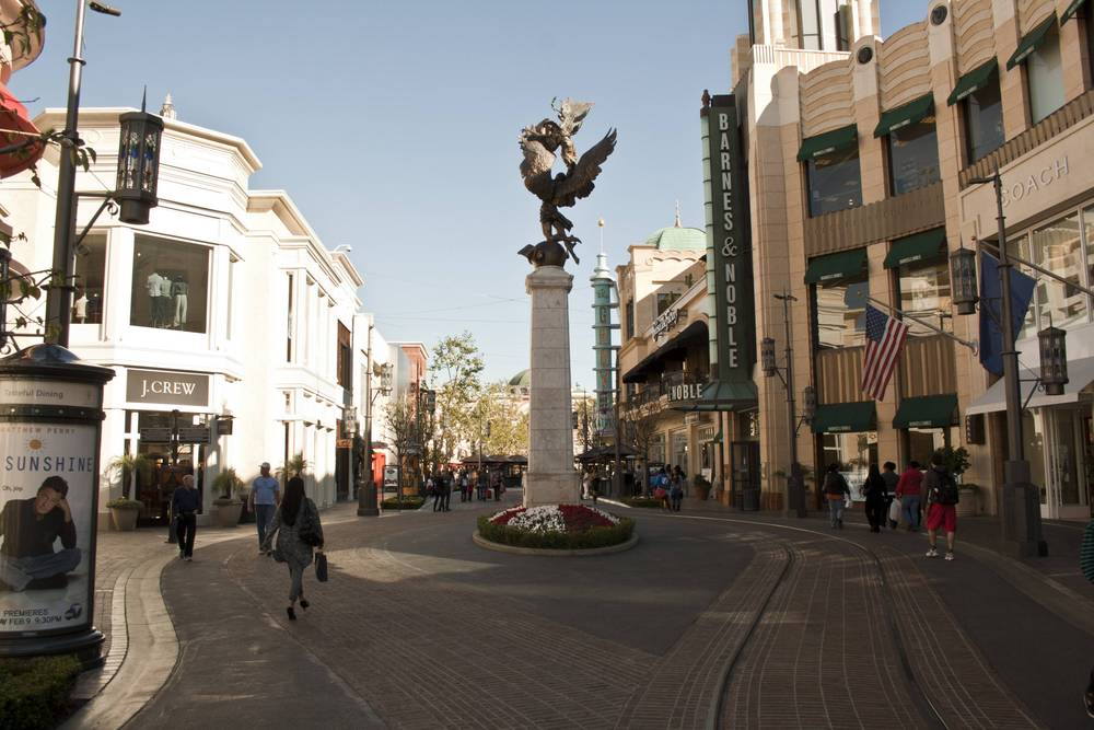 Shoppers walk past upscale storefronts along a pedestrian street with a fountain statue.