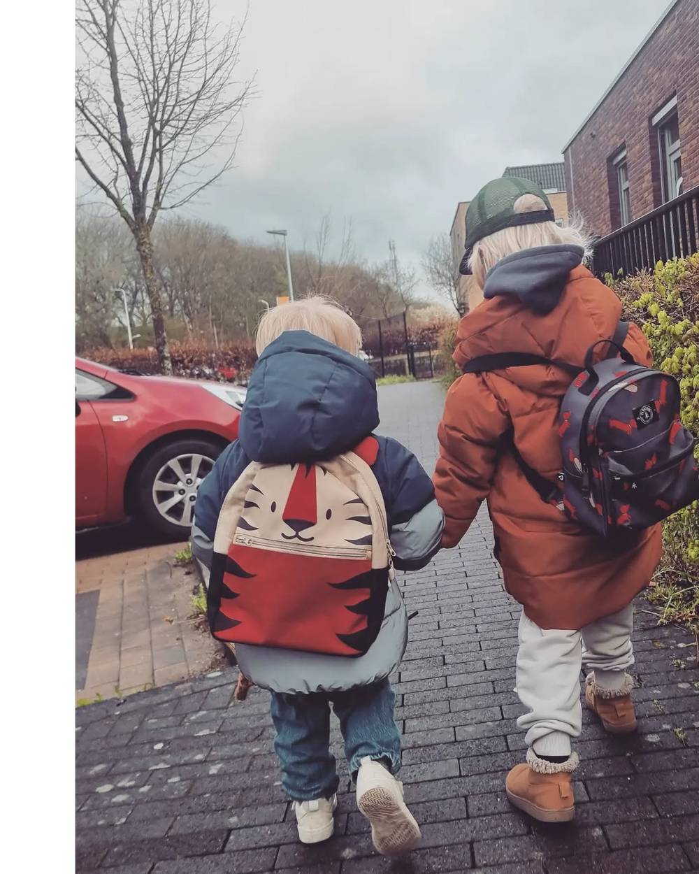 Two children walking hand in hand down a residential street wearing backpacks.