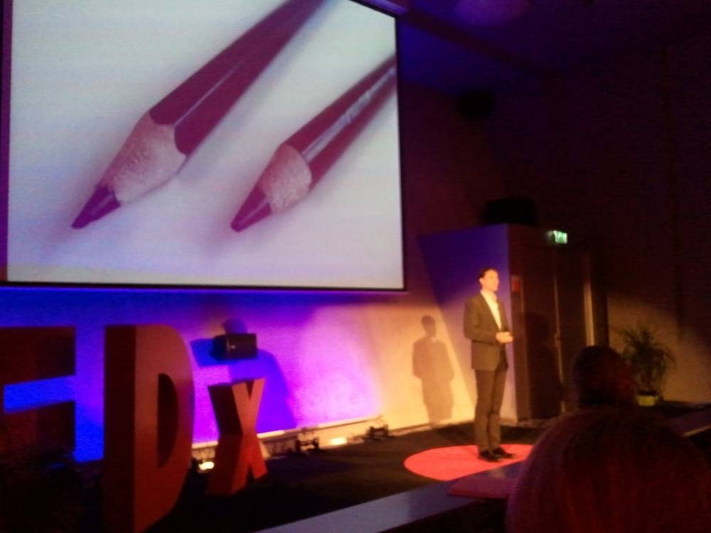 A speaker stands on the red TEDx stage in front of a slide showing two pencils.