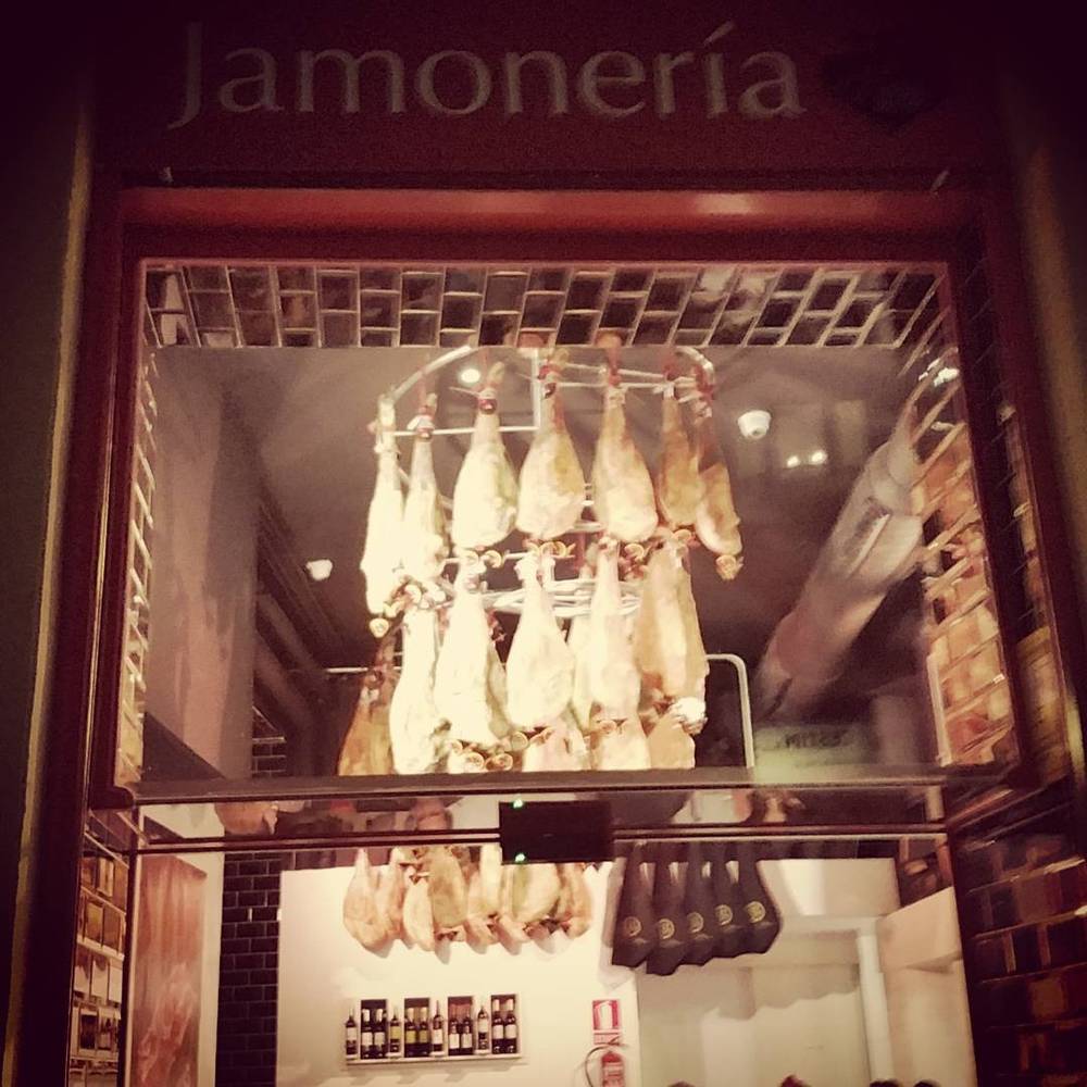Cured hams hanging in the window of a jamoneria (ham shop) at night.