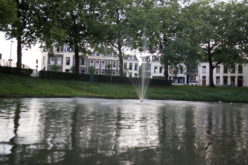 A water fountain jetting up from a canal lined with historic townhouses and trees at dusk.