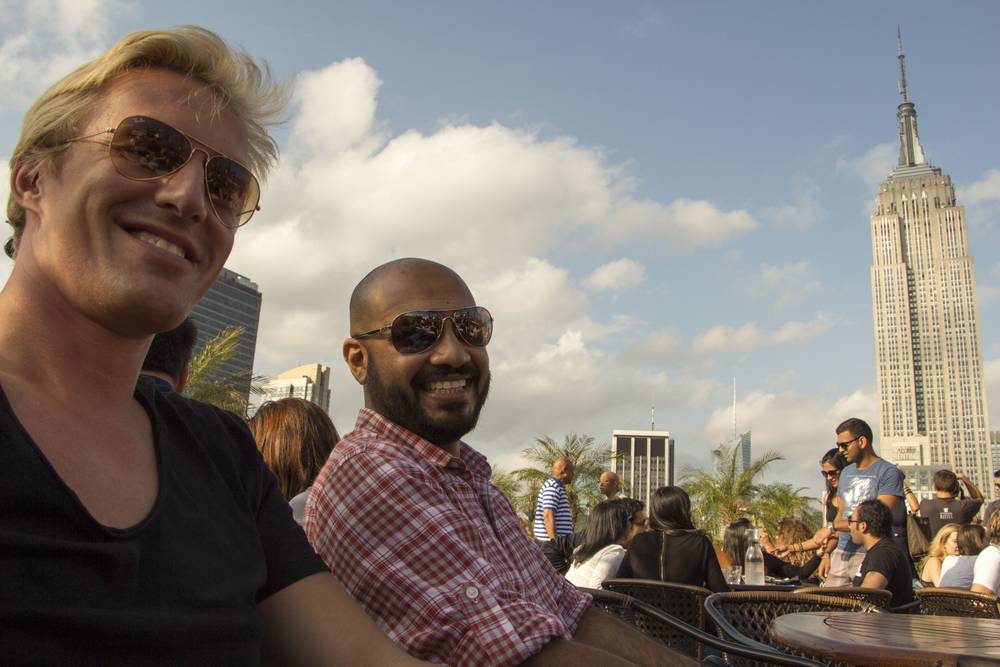 Two men smile at a rooftop bar with the Empire State Building behind them.
