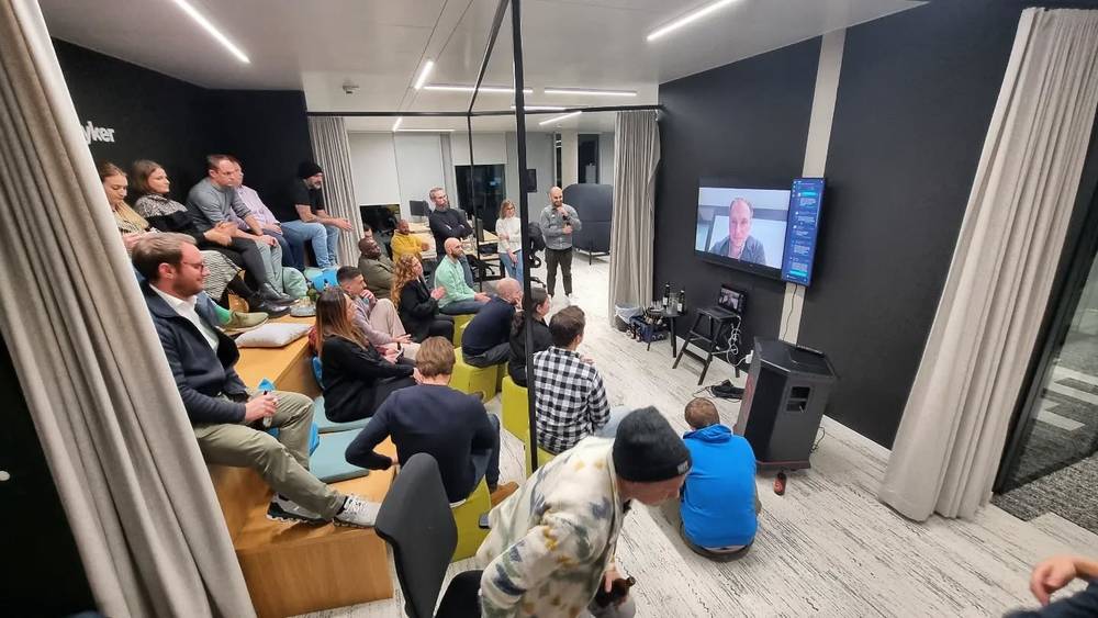 An audience seated in a modern office lounge watching a speaker appear on a wall-mounted screen.