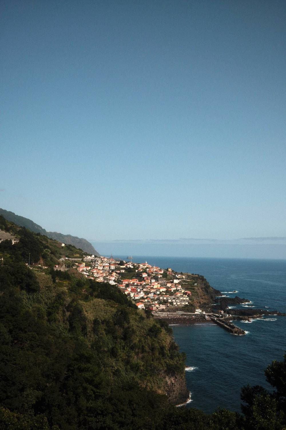 A coastal village with terracotta-roofed houses cascades down a steep green hillside toward the Atlantic Ocean on the island of Madeira, Portugal.