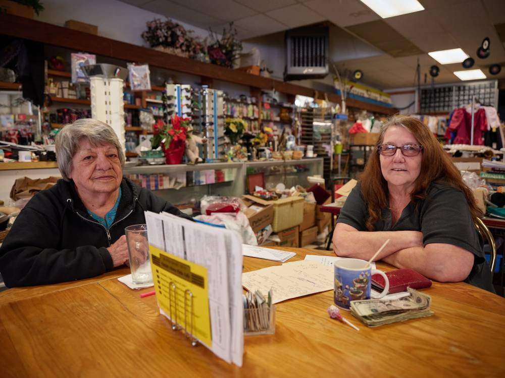 Restaurant owner Pattie Deitz sits alongside her best friend Jenny at a worn wooden table in Pattie's Home Plate Café in Portland's Saint Johns neighborhood. The intimate portrait captures the warmth of longtime friendship against the bustling backdrop of the eclectic eatery, where cluttered shelves overflow with collectibles, flowers, and memorabilia. Soft fluorescent lighting bathes the scene as the two women pause from their daily routines, newspapers and coffee cups scattered before them.