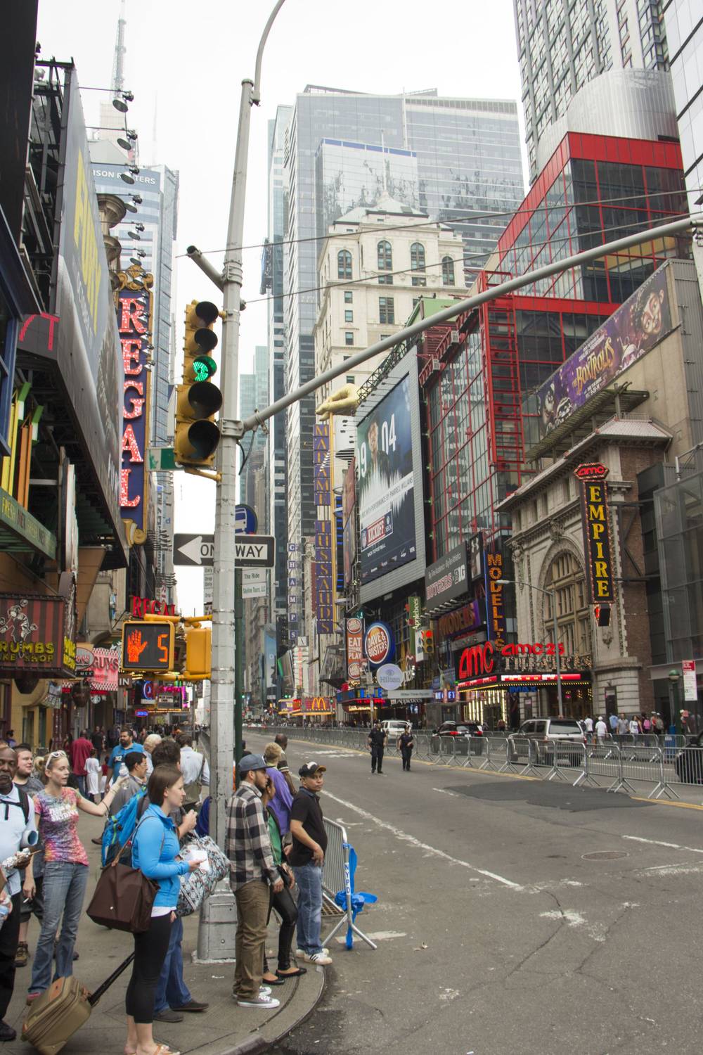 Pedestrians wait at a barricaded Times Square corner beneath theater marquees.
