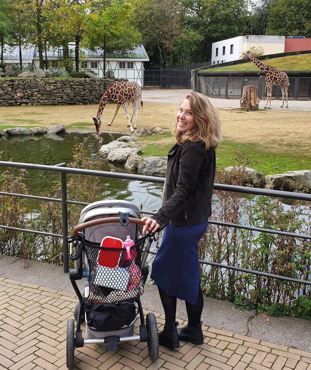 A woman pushing a stroller past a giraffe enclosure at a zoo.