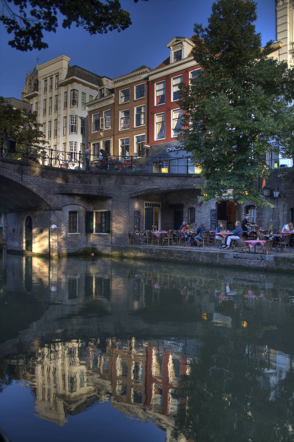 Historic canal-side buildings and an outdoor cafe reflected in the still water of the Oudegracht canal at dusk.