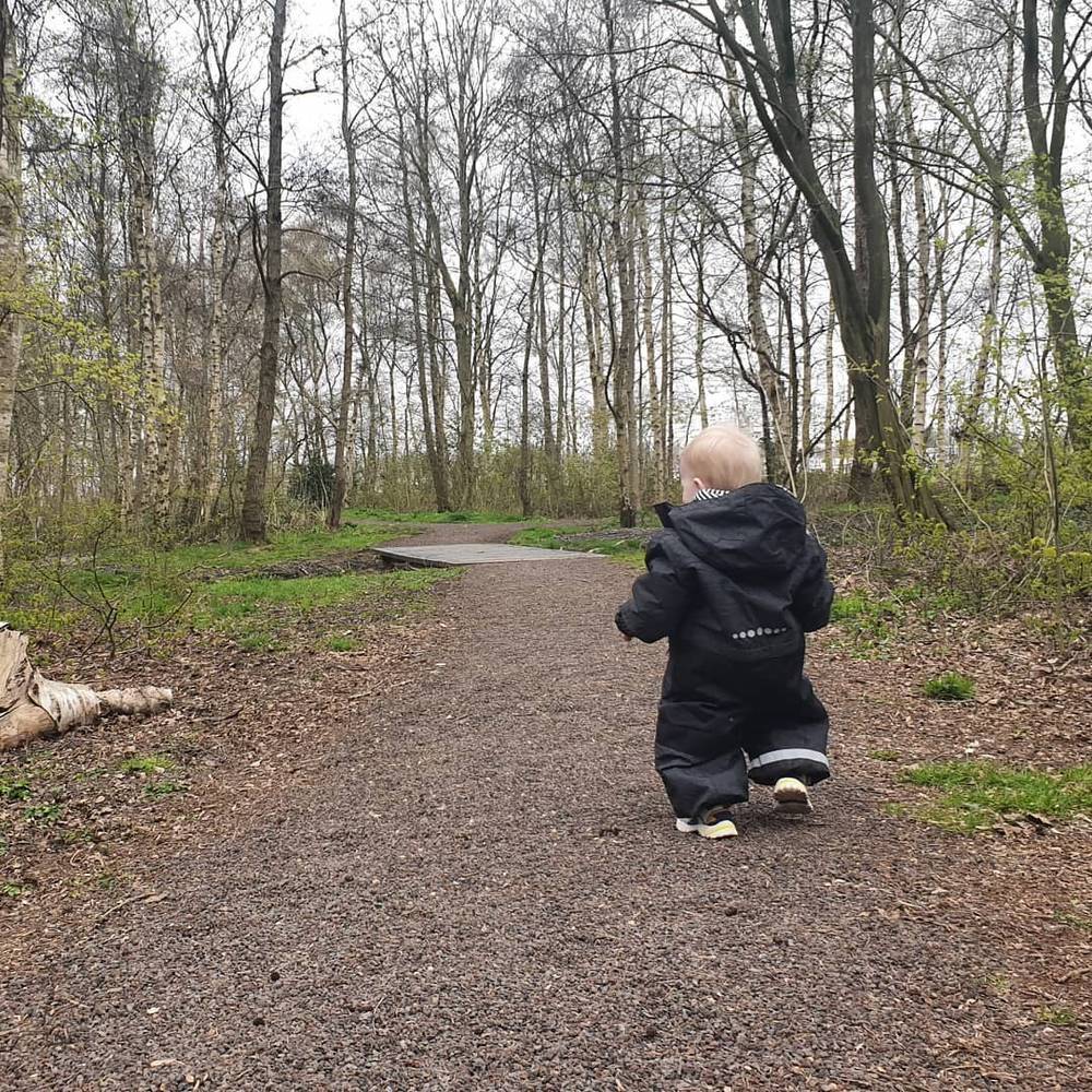 A child in a dark rain suit walking alone along a wooded gravel path.