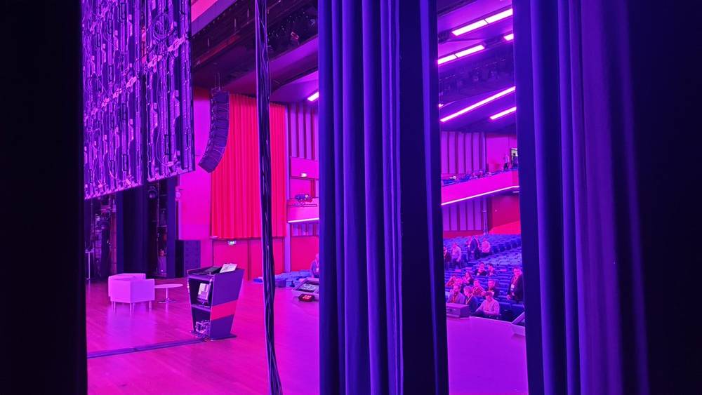 A wide view of an empty theatre stage lit in purple with red curtains and balcony seating.