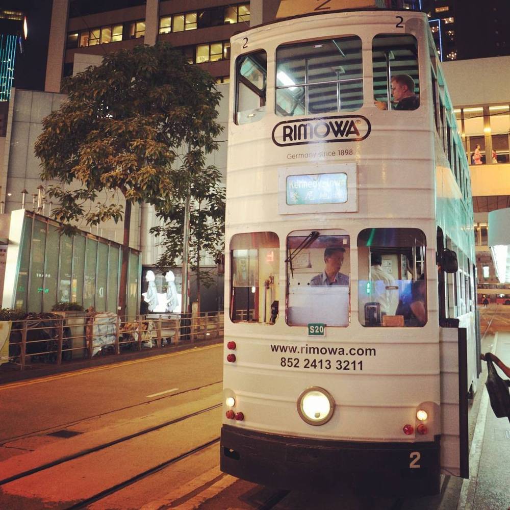 A white double-decker tram with a Rimowa advertisement driving through a Hong Kong street at night.