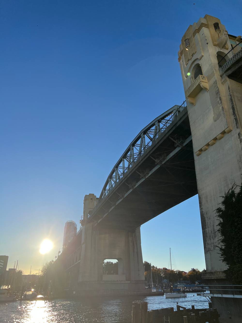 Burrard Bridge from underneath with boats docked below and the sun low in the clear sky.