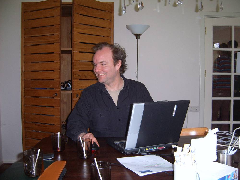 A man smiles at a laptop at a wooden table, louvered shutters framing the window behind him.