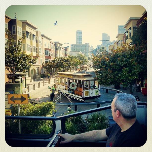 A man rides in an open-top tour trolley in a city street lined with buildings and a cable car.