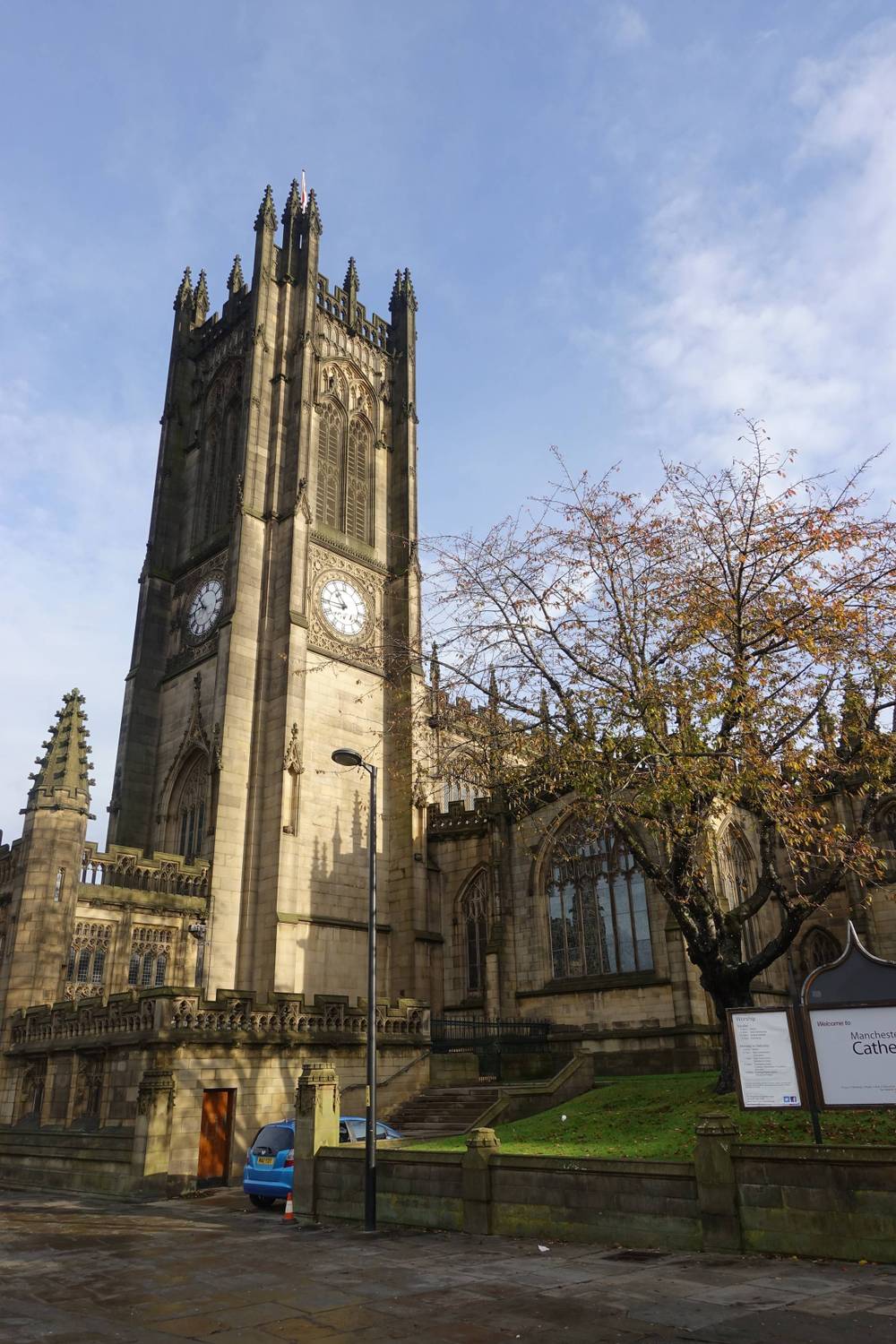 Manchester Cathedral's square clock tower stands beside autumn trees.