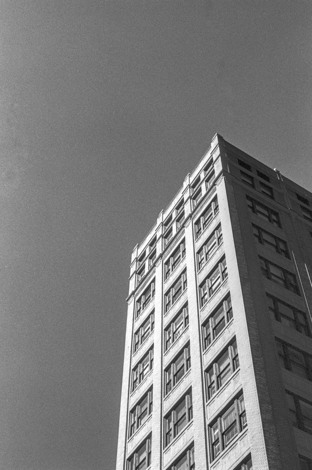 Low-angle view up the corner of a slender pre-war commercial tower. Buff brick walls with stone-trimmed paired windows rise in a tight rhythm against a clear sky. The right face recedes into shadow; the left face is well lit.