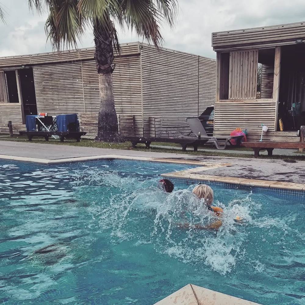 Two people swimming and splashing together in a pool beside wooden cabin-style buildings.