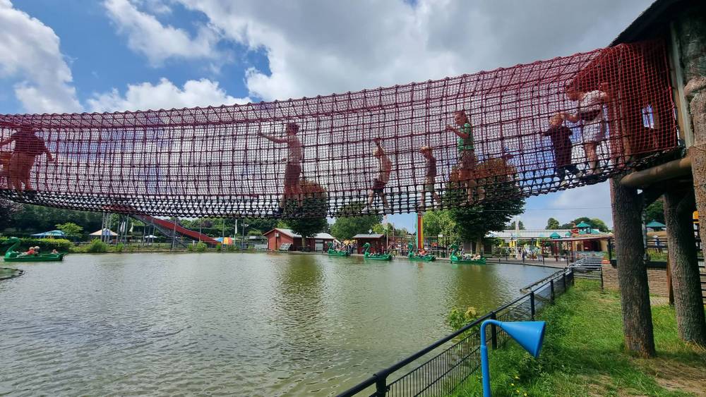 Children climb a red net-covered rope bridge structure over a pond.