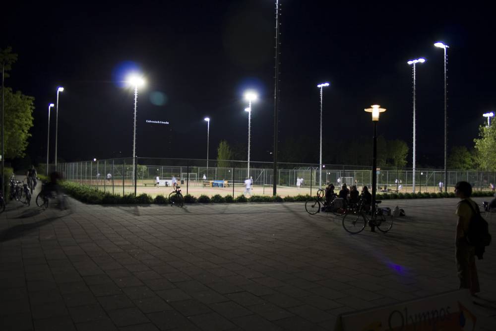 Floodlit sports courts are busy with people and parked bicycles at night in Utrecht.