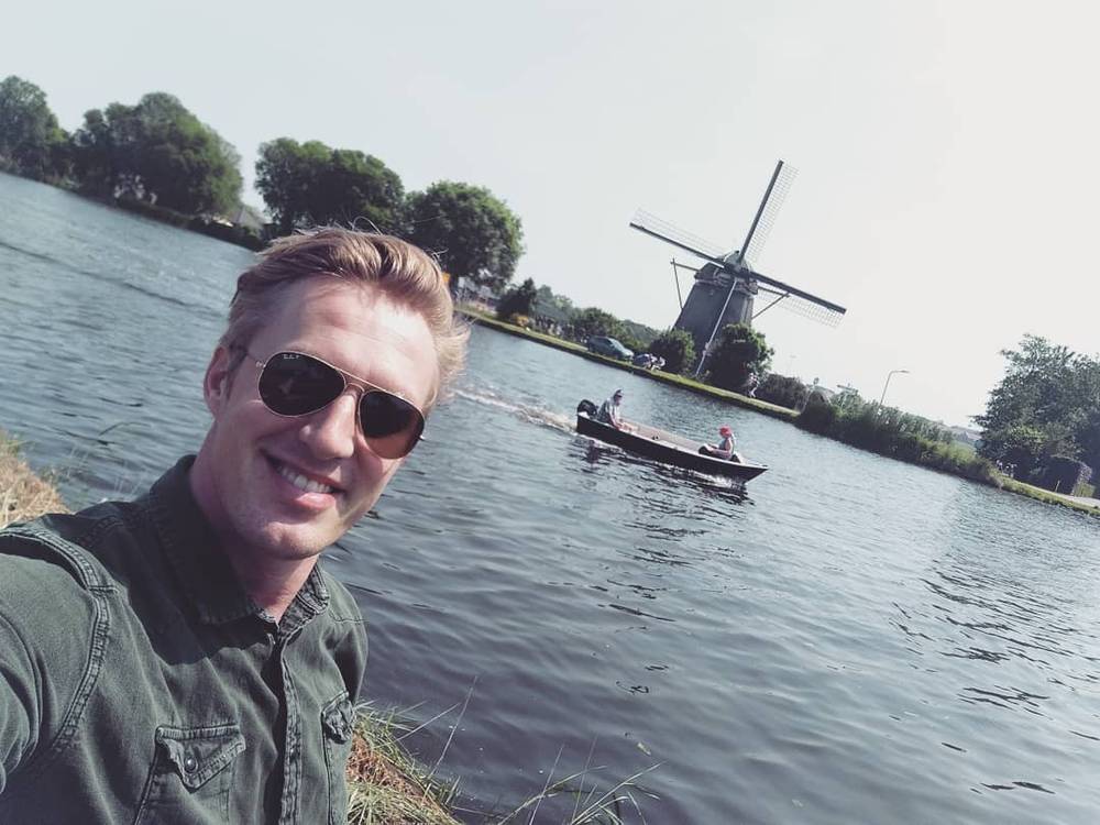 Selfie by a river with a traditional Dutch windmill and a rowboat in the background.
