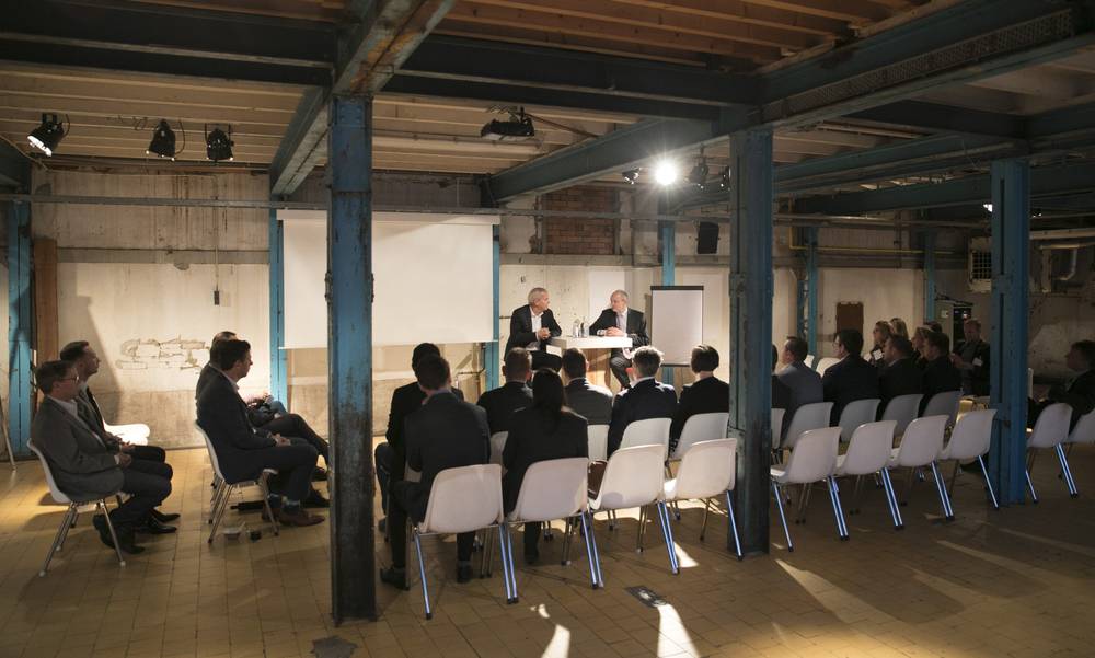Rows of people sit facing a small stage and projector screen under a railway arch.