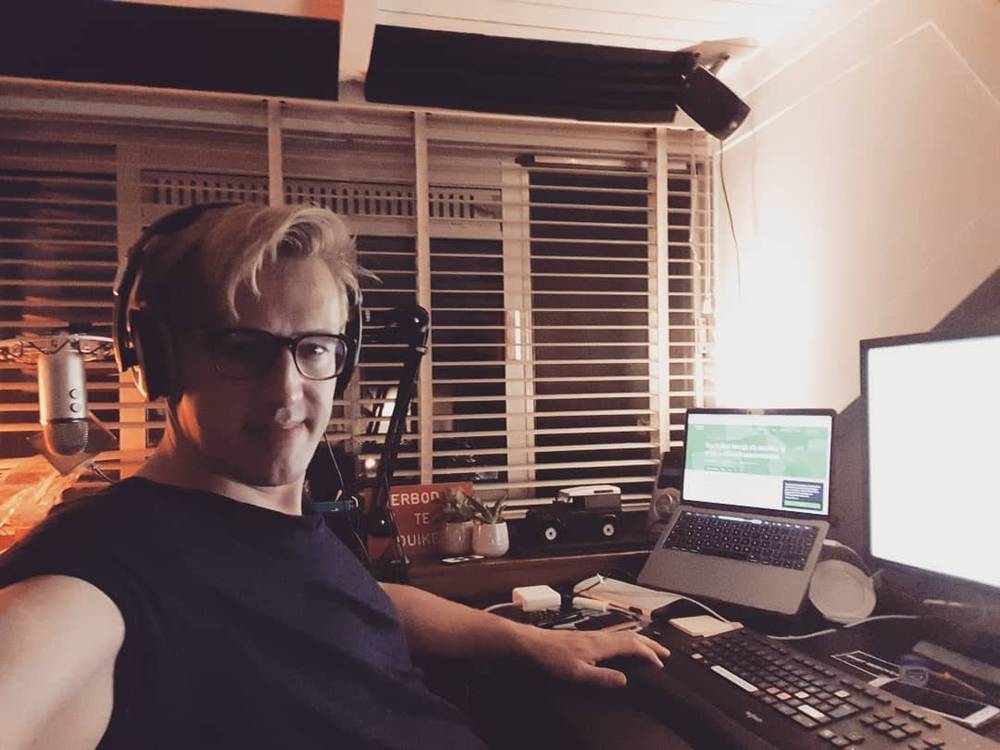 Selfie wearing headphones at a desk with a laptop and monitor, in a wood-paneled room.