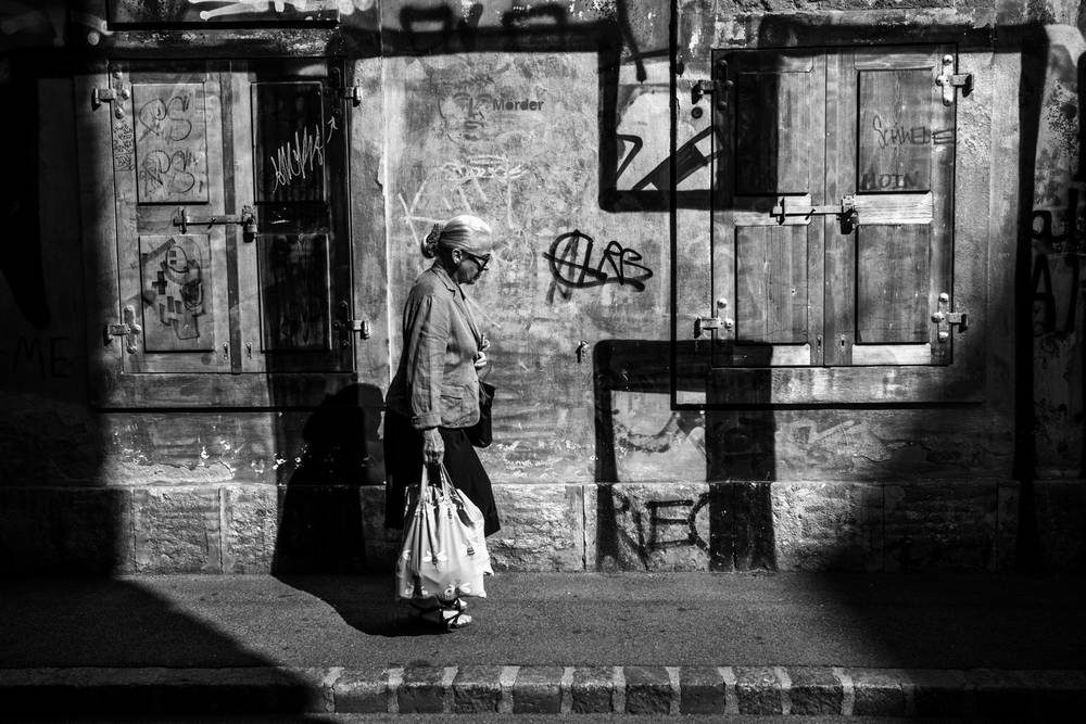 ⁠A high-contrast black and white photograph of an elderly woman with white hair, viewed from behind and the side. She is walking on a city sidewalk and carrying a white plastic shopping bag. Her shadow is long. The background is a textured, weathered concrete wall with a closed wooden shutter-door and extensive, distinct graffiti, including stylized tags and lines. The lighting creates a strong dramatic contrast between deep shadow and highlighted elements. The image is presented with a white border.