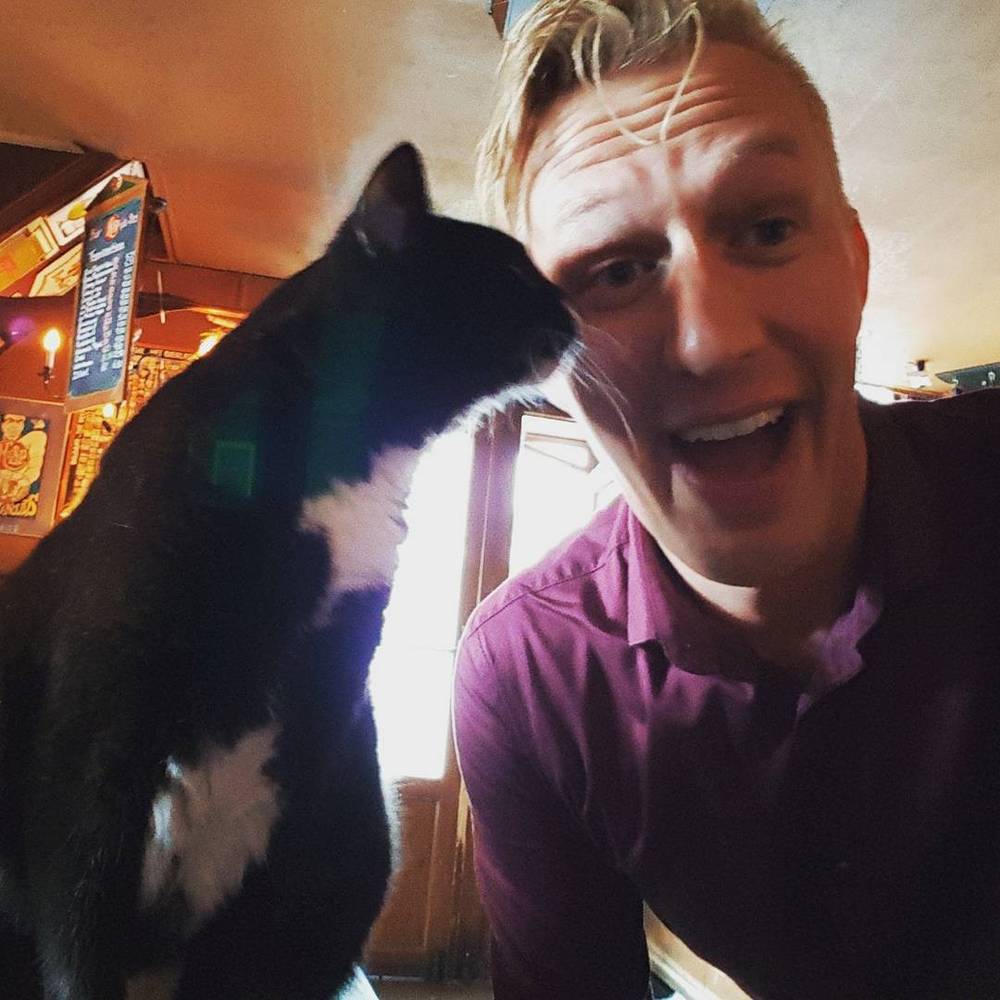 Close-up selfie of a man with a black and white cat nuzzling his face.