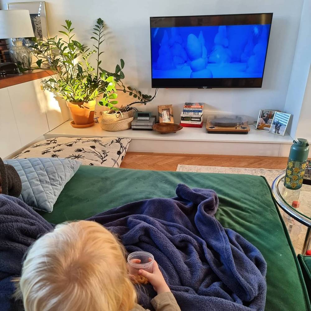 A cozy living room with a TV on the wall and a person under a blanket on the sofa holding a drink.