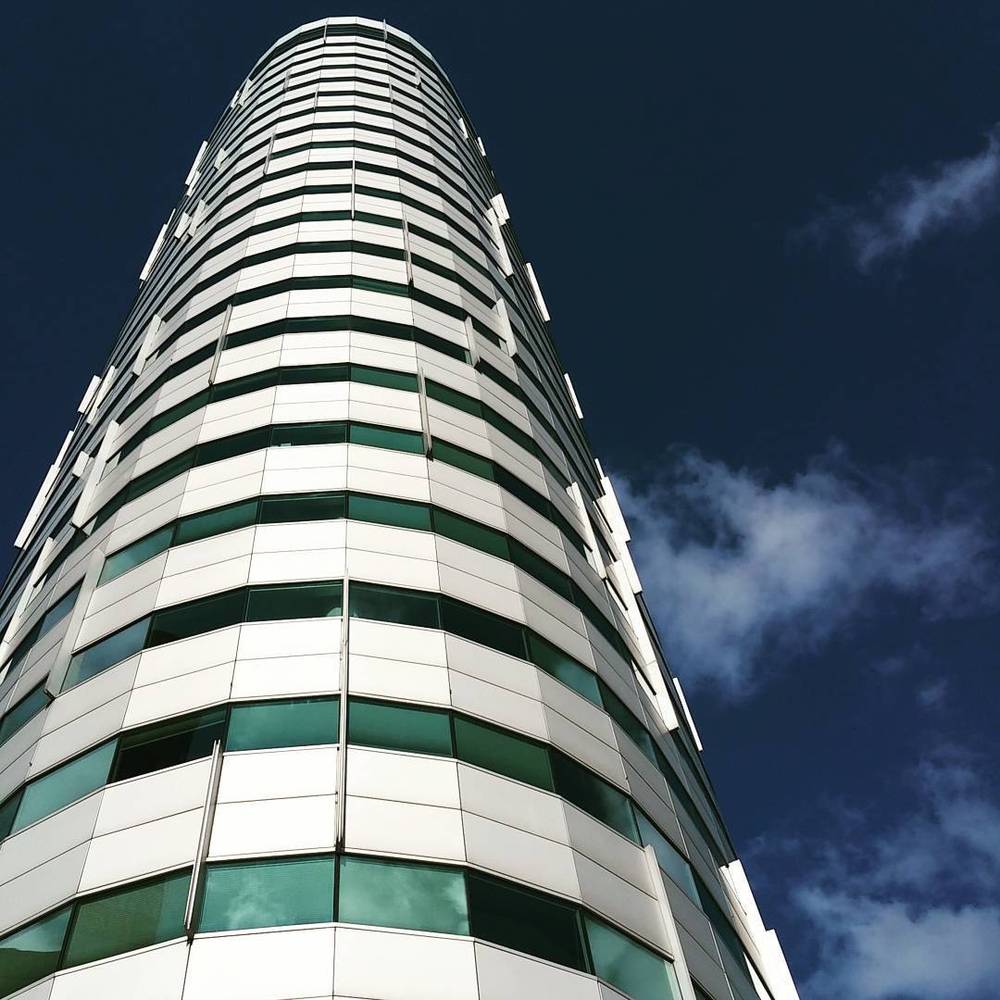 Looking straight up at a curved high-rise building with alternating white and dark green glass panels against a blue sky.