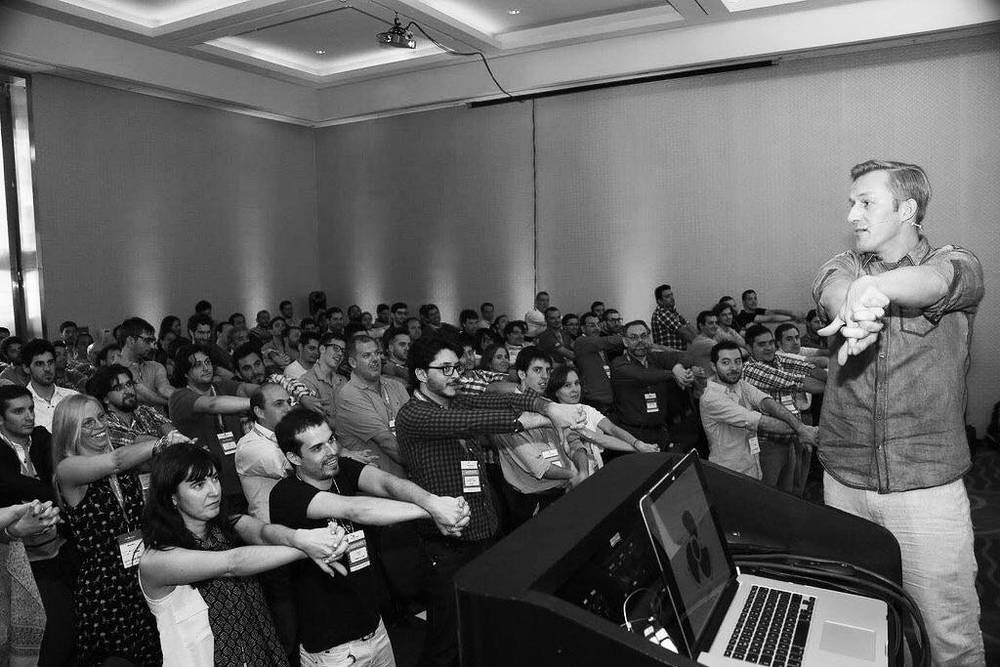 A black and white photo of a speaker leading an audience in synchronized arm movements at a conference.