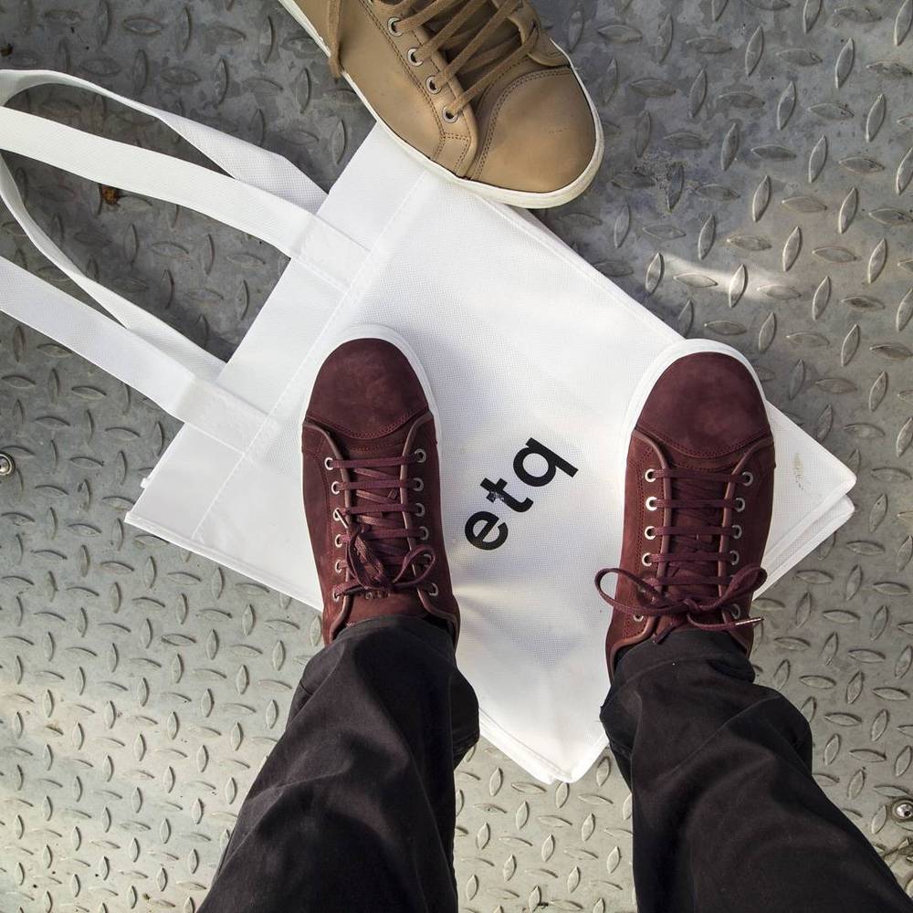 A canvas tote bag reading "etq" beside a pair of brown sneakers on a metal floor.