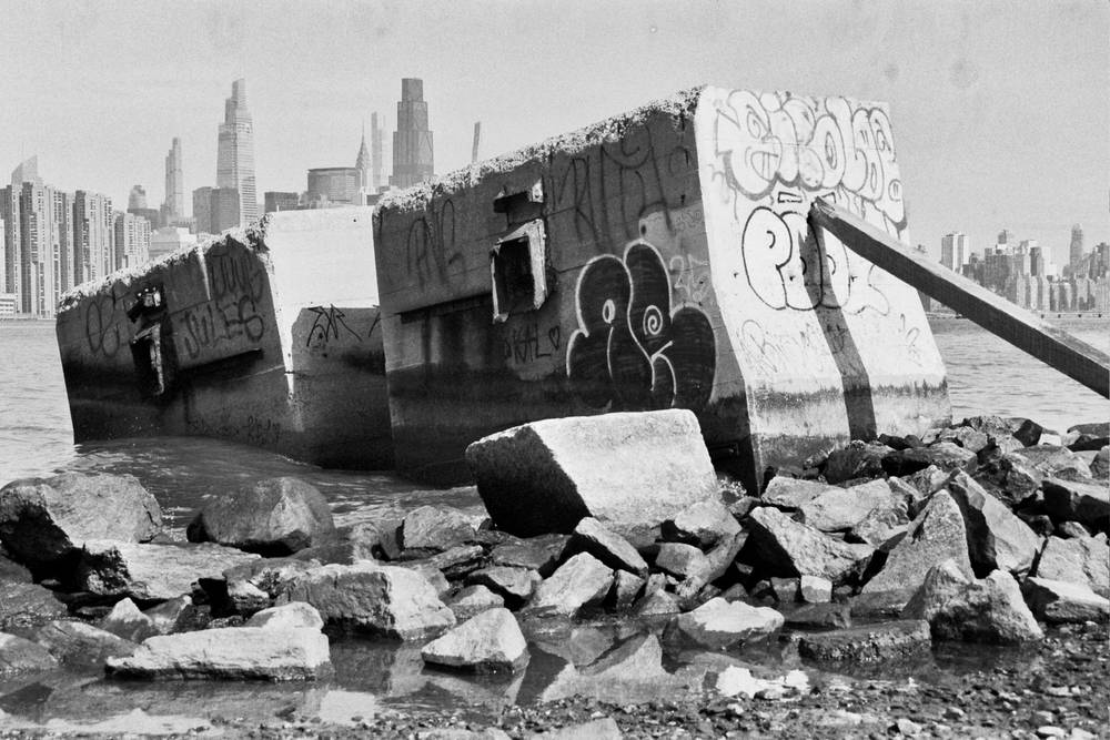 Two massive concrete blocks sticking out of the water near the Brooklyn shore of the East River. The one further away  is lower and both are at an angle, appearing to be sinking slowly. The Manhattan skyline is visible in the distance. Black and white.