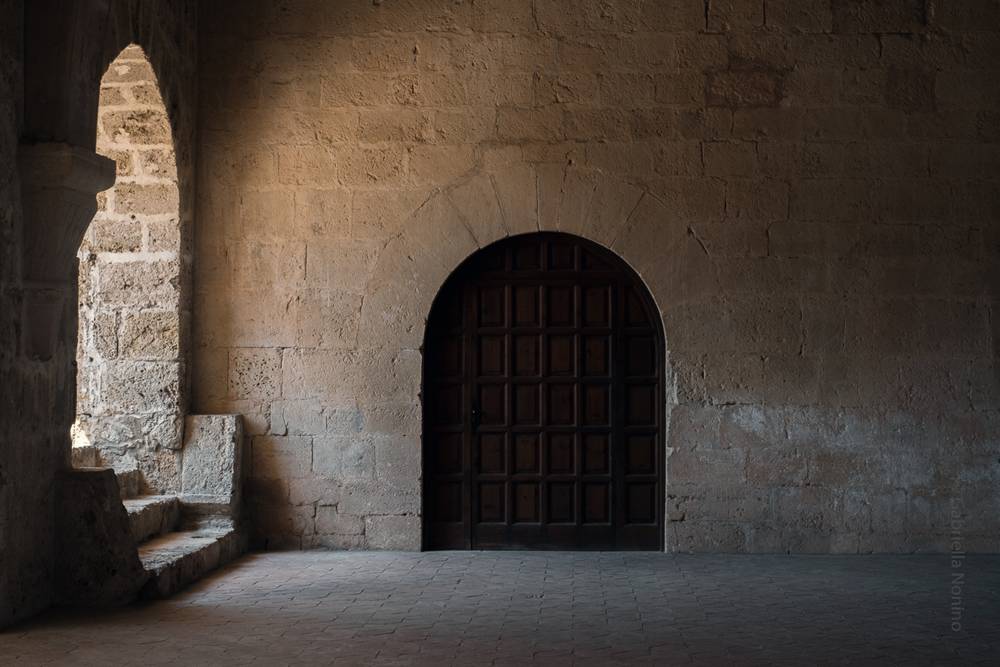 Dark passageway wall with an arched wooden door and a stepped stone bench beside a narrow lit archway to the left.
