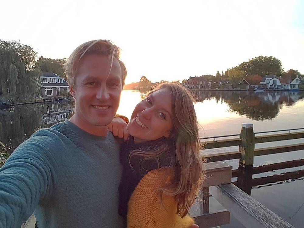 Selfie of a couple smiling together at sunset by a lake, with houses reflected in the water.