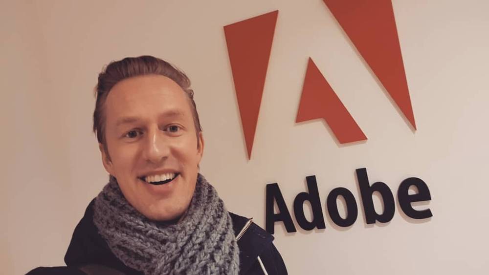 A man smiles in front of a large Adobe logo sign.
