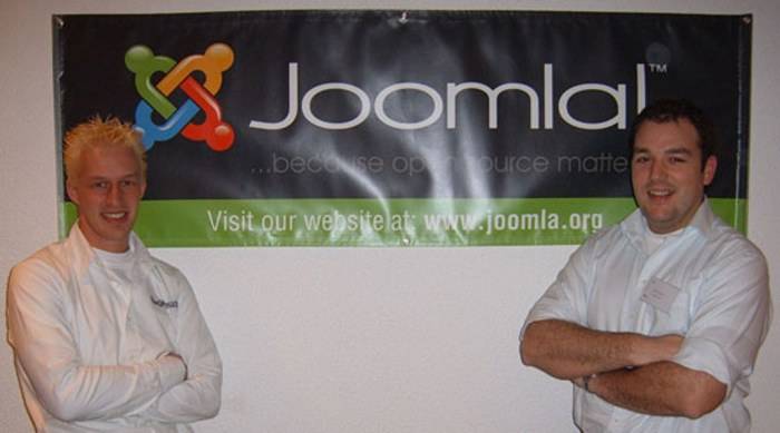 Two men in white shirts pose crossed-armed in front of a Joomla banner.