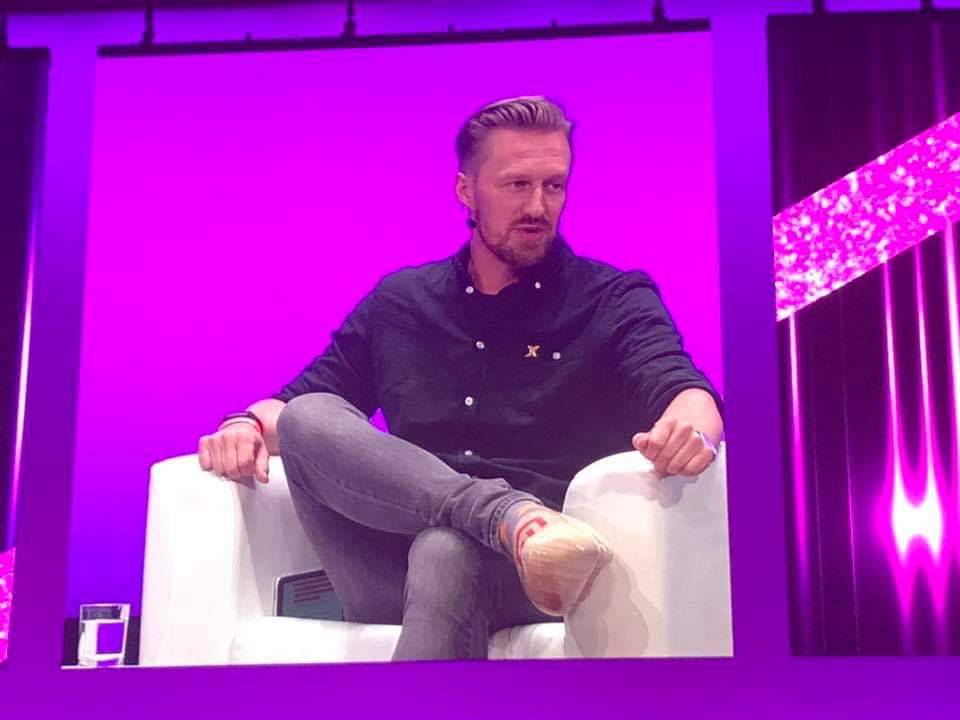 A man sits casually in a white chair on a purple lit stage during an interview segment.