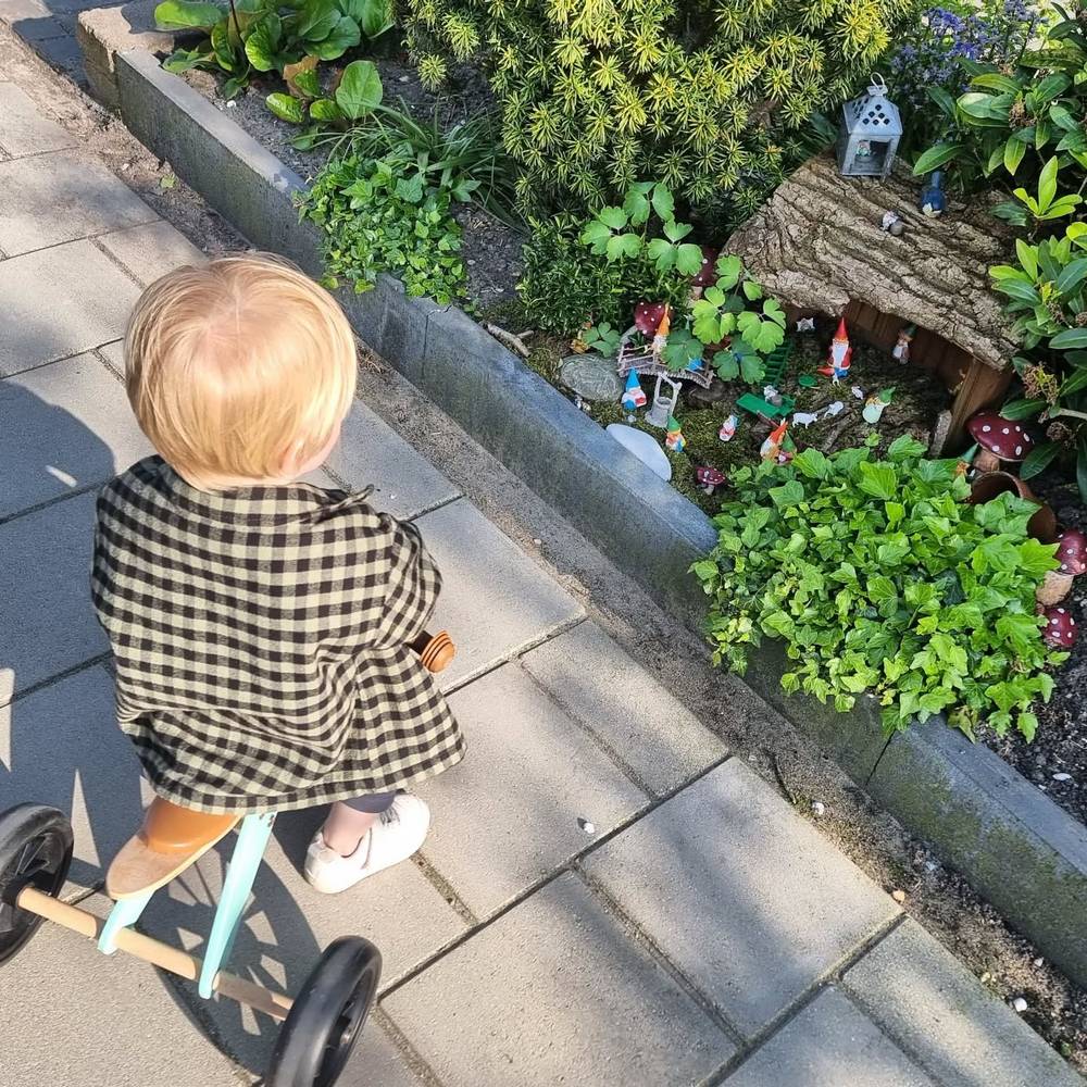 A toddler on a small balance bike looking at a miniature garden gnome village along a garden path.
