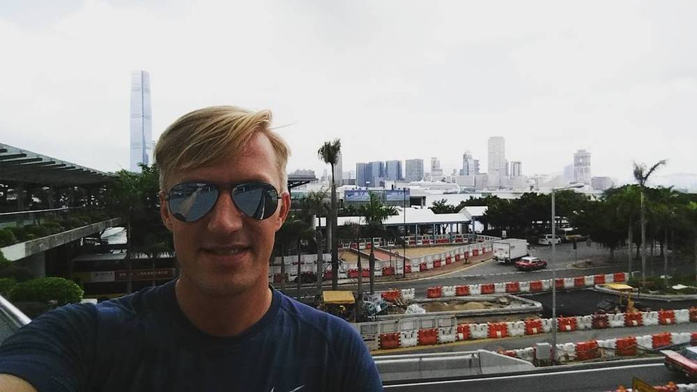 A selfie of a man in sunglasses with the Hong Kong skyline and palm trees behind him.