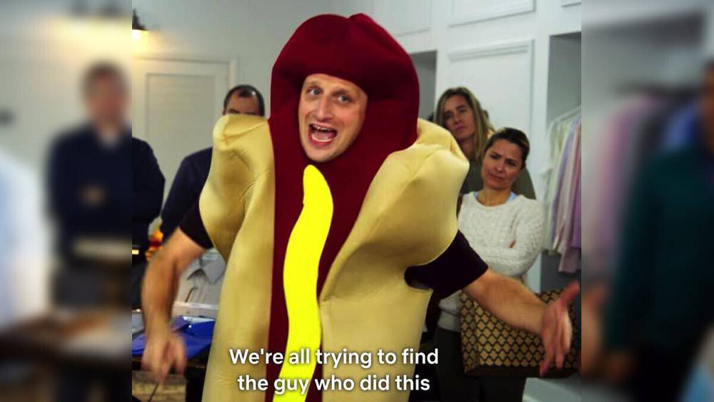A person dressed in a hot dog costume is speaking animatedly in a room with several people watching, accompanied by the caption We’re all trying to find the guy who did this.