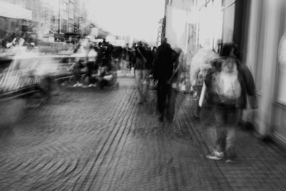 Black and white motion blur photograph of people walking along a busy city street, creating a dynamic and ethereal sense of movement.