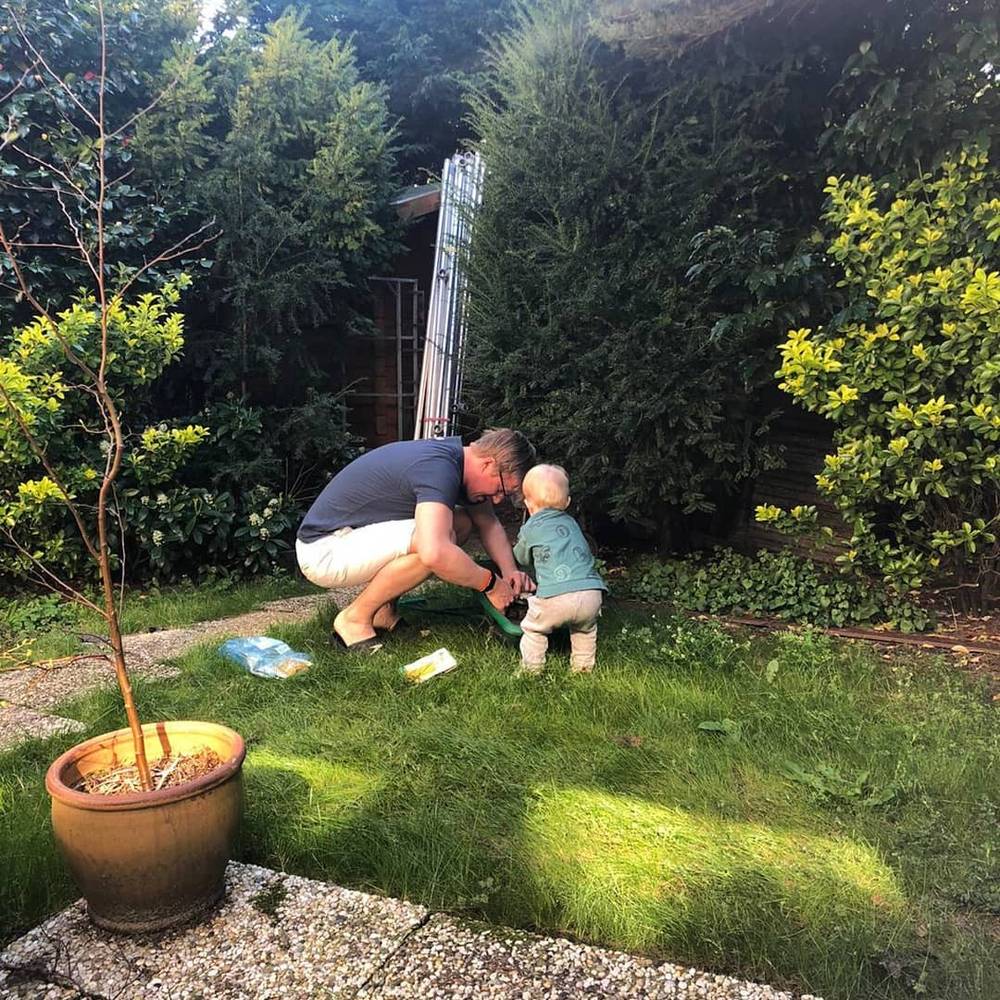 A man crouches in a garden helping a toddler plant something in the grass, with a potted young tree nearby.