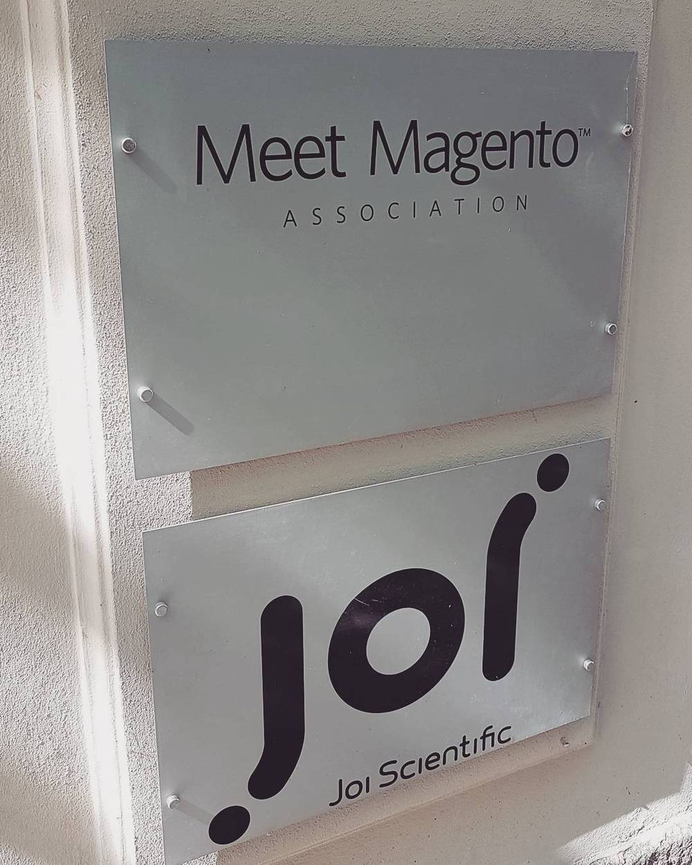 A wall-mounted sign reading "Meet Magento Association" above a "Joi Scientific" plaque.