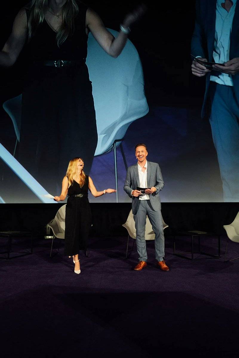 Two people standing on stage, one gesturing, at an evening conference event.