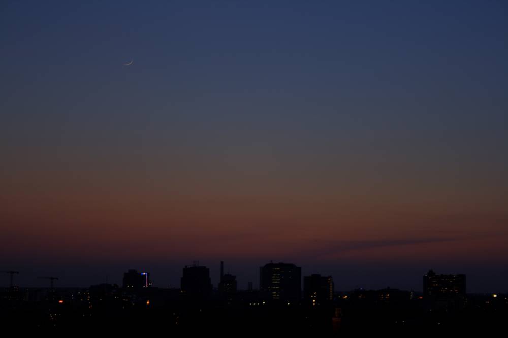 A crescent moon hangs in the darkening sky over the Utrecht skyline at dusk.